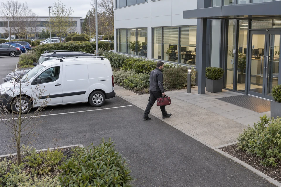 A contractor carrying a burgundy tool bag from a van towards an office entrance.