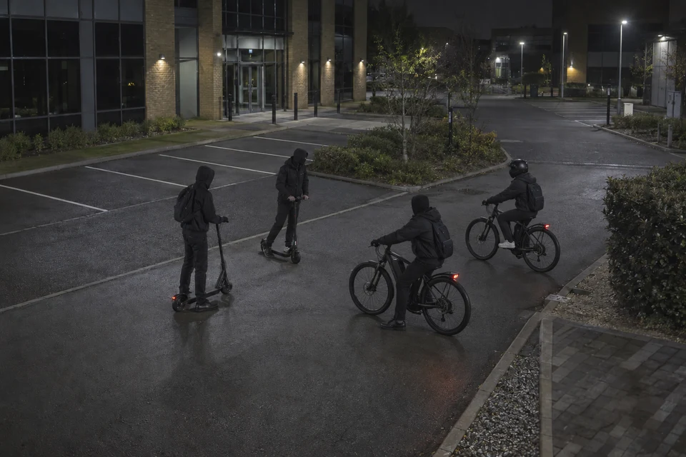 Four people with covered faces on two electric scooters and two electric bicycles outside closed offices at night.