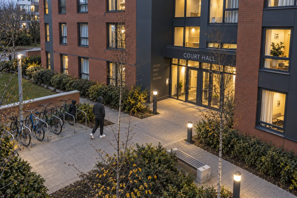 A person in a charcoal hoodie, black trousers and white trainers approaching a student residence at dusk, without a backpack.