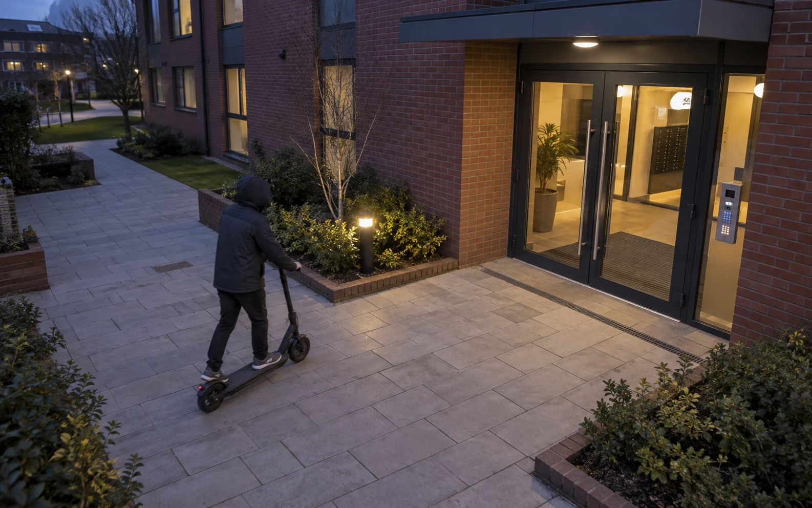 Elevated view of a hooded adult arriving on an electric scooter outside a student accommodation entrance at dusk.